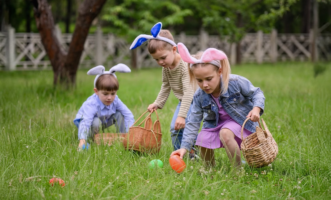 three children wearing bunny ears collecting colorful easter eggs in baskets during outdoor easter egg hunt in green meadow